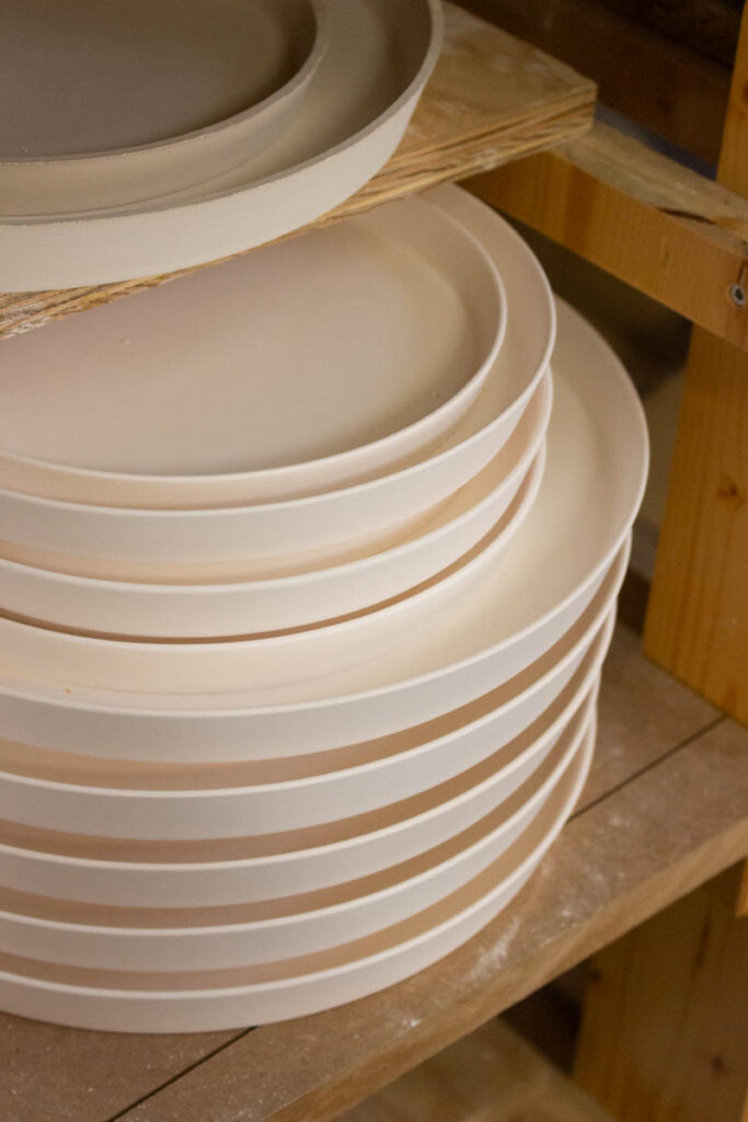 Close-up of stacked unglazed bisque porcelain plates on wooden shelving at the studio